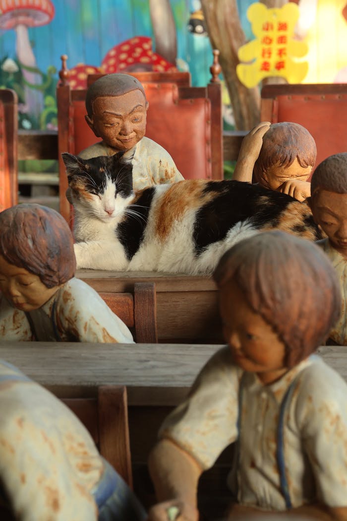 A serene calico cat lying on a wooden desk surrounded by child sculptures.